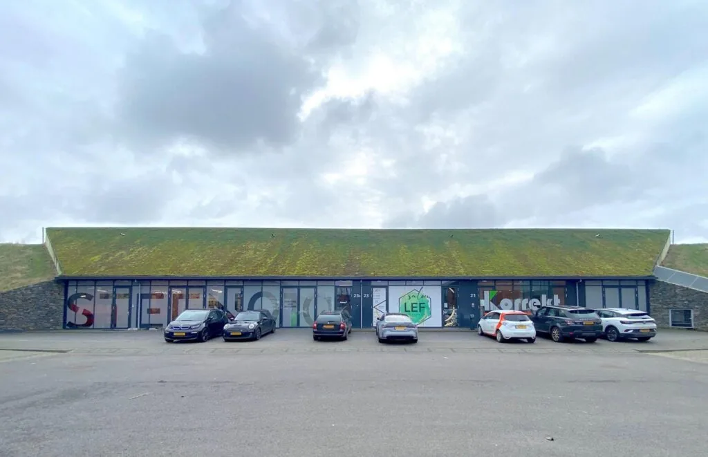 Moderne bedrijfsruimte met een groen begroeid dak en geparkeerde auto’s voor de gevel, waaronder Salon LEF en Korrekt.